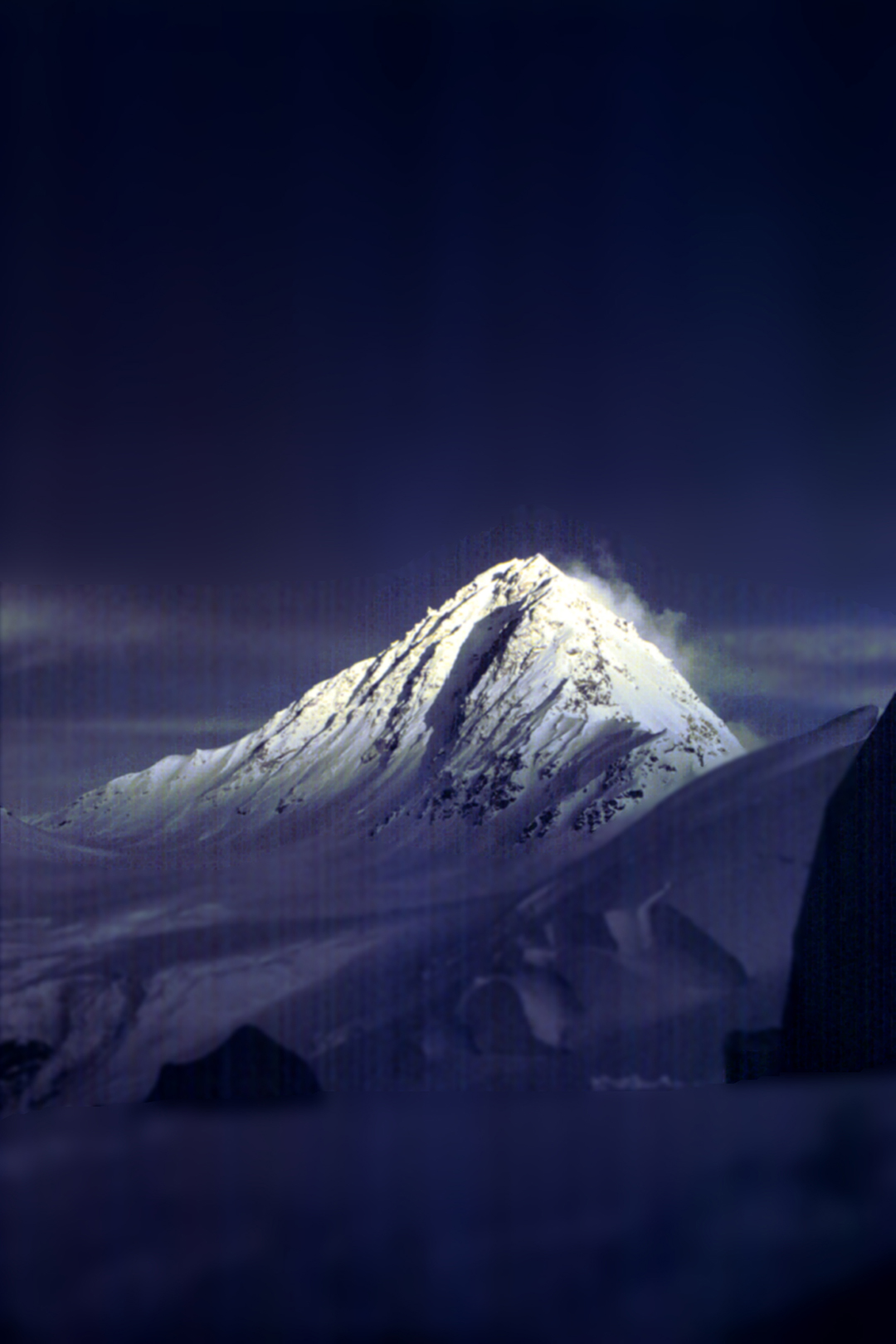 A snow covered mountain peak lit up by natural light under dark blue skies. 