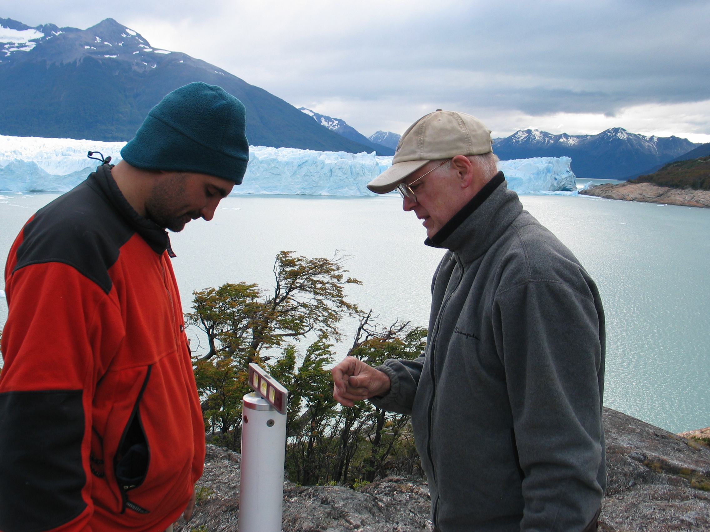 men looking at equipment in front of a lake and snow capped mountains  