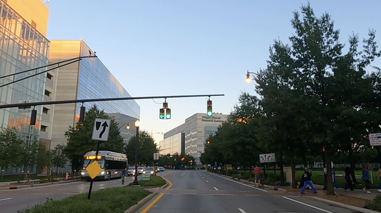 A street view image of green traffic lights, with sidewalk, trees, buildings, bus and cars  on the left, building straight ahead  and green grass median and park, sidewalks and trees and people walking