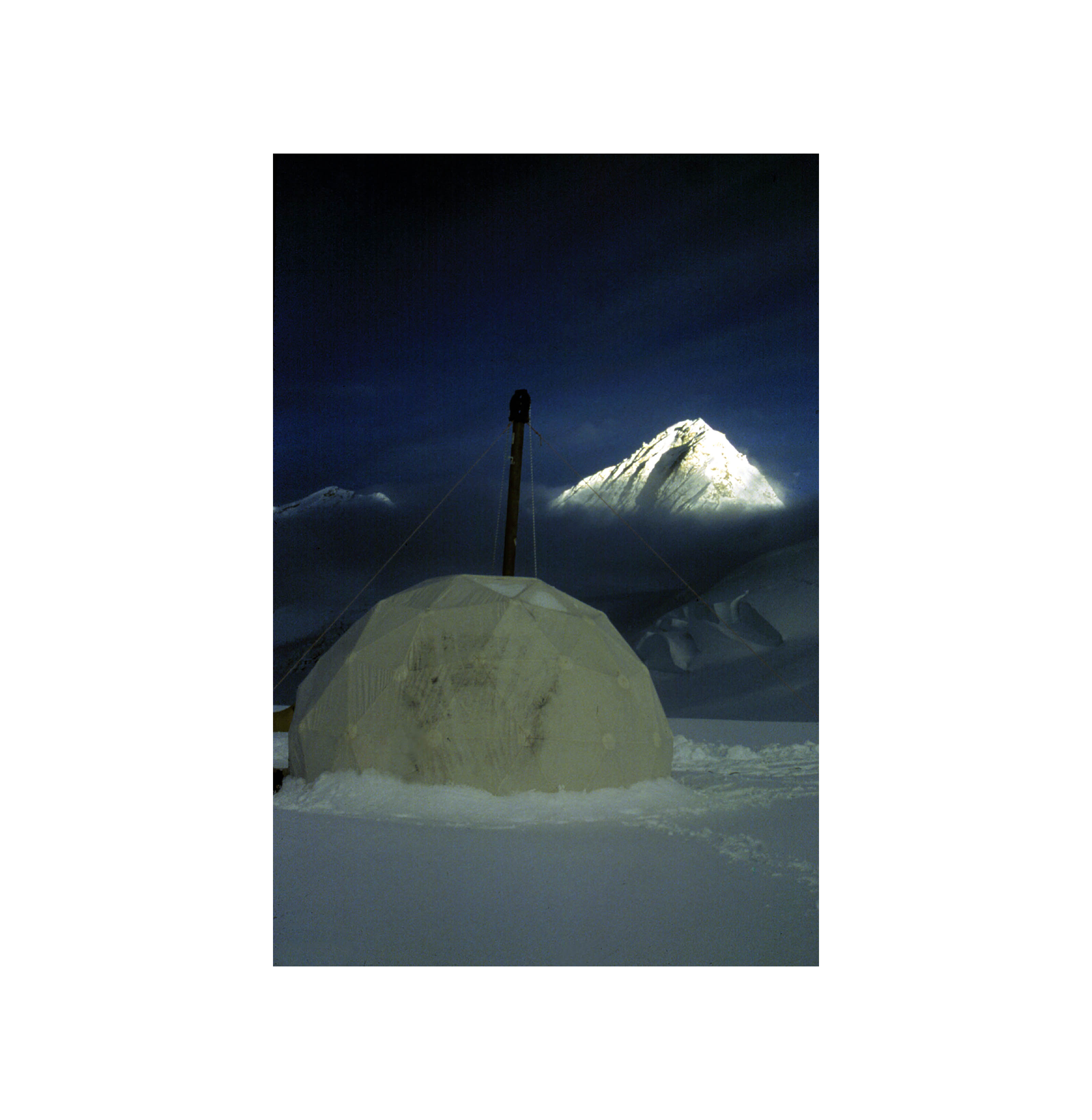  A white dome tent on the snowy ground and a white snow capped mountain at a distance under dark blue skies.