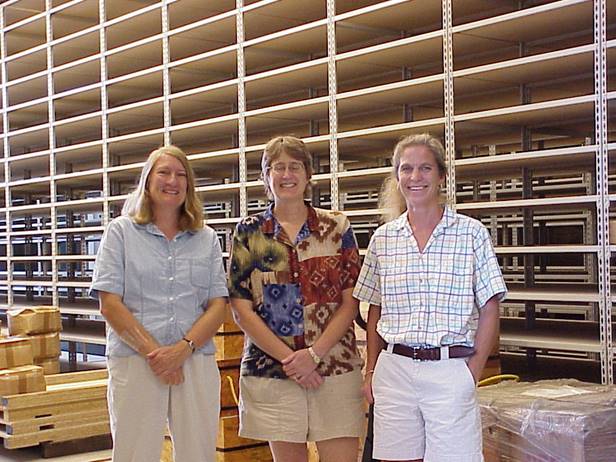 Three women standing in a room with what appears to be floor to ceiling empty, wooden shelving behind then. 