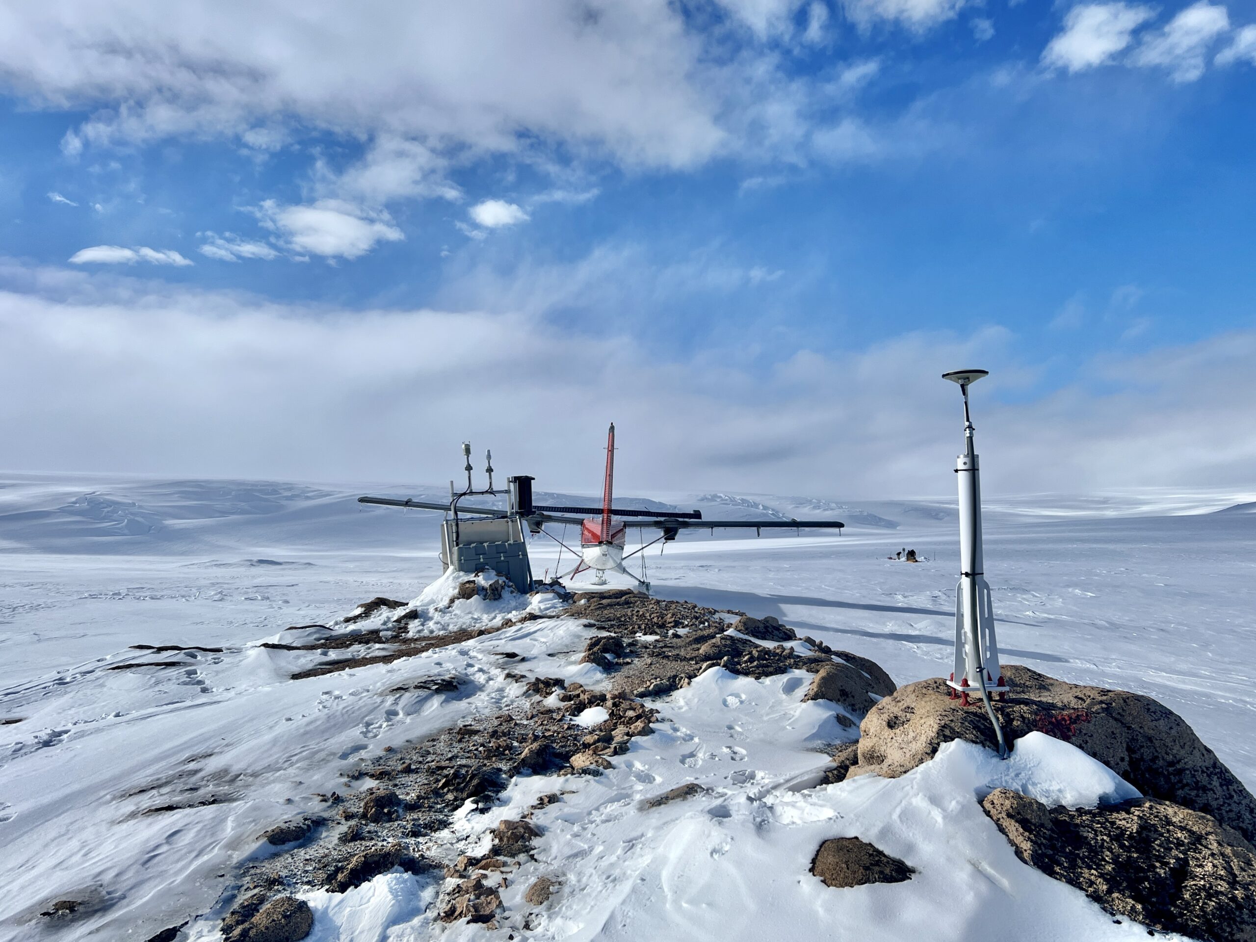 A vast snowy field with some rocks, equipment and a plane in the foreground under blue skies with white clouds.