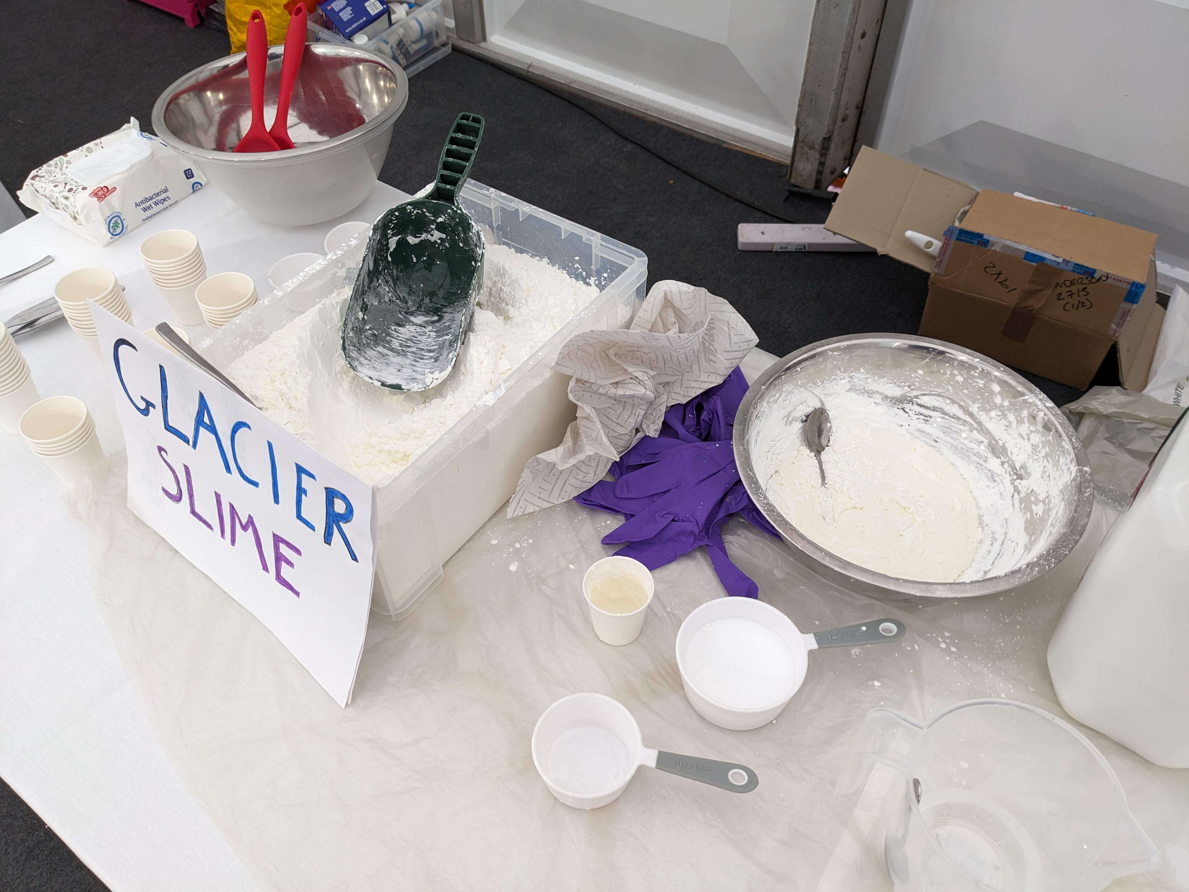Ingredients on a table, including white powder, measuring cups, bowls and a sign that says Glacier Slime. 