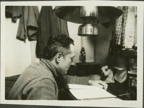 a man reading at a desk in a room with a light fixture above his head and a small dog laying on the desk in front of him.