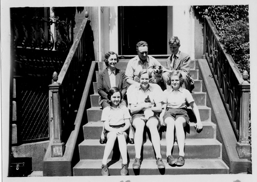 Black and white of a family of six sitting in two rows of 3, on front steps of a building with pets.