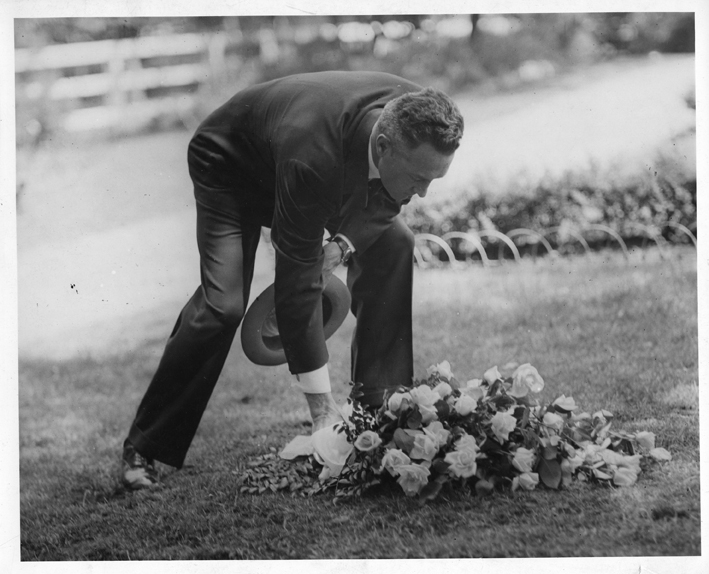 A black and white photo of a man bent over to add to the flowers covering a gravesite.