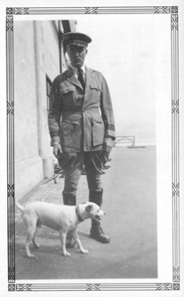 Black and white image of a man in uniform standing with a dog in front of him.