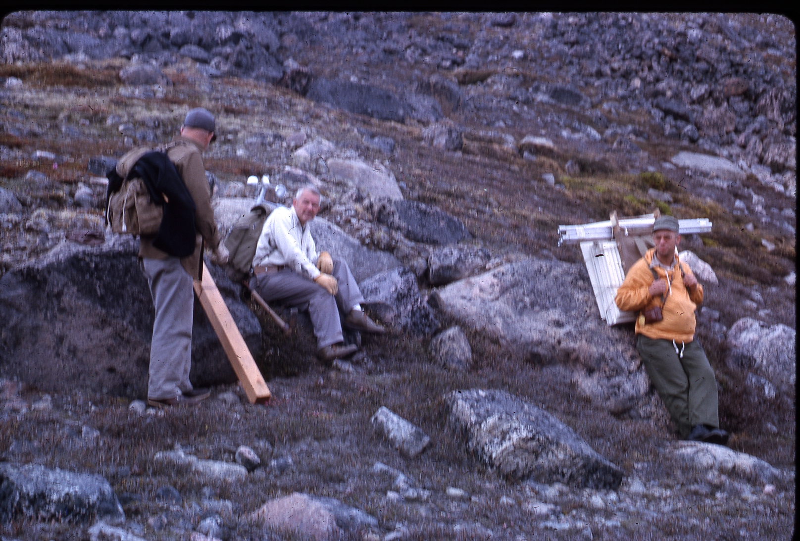 Three people in a rocky mountainous field raking a break, one sitting and one leaning on rocks.