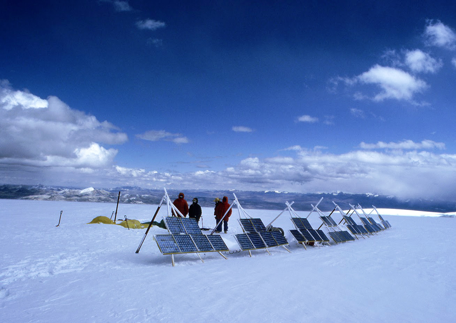 People standing behind a row of solar panels installed on easels on a snowy ground, surrounded by mountains in a distance under blue skies and white clouds.  