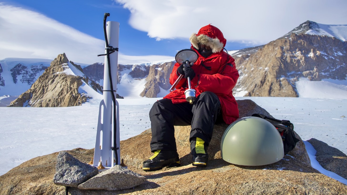 A person sitting on a bedrock with a red ski jacket, seated in front of the GNSS antenna, holding an object. In the background are mountains and snow covering some of them and the ground.