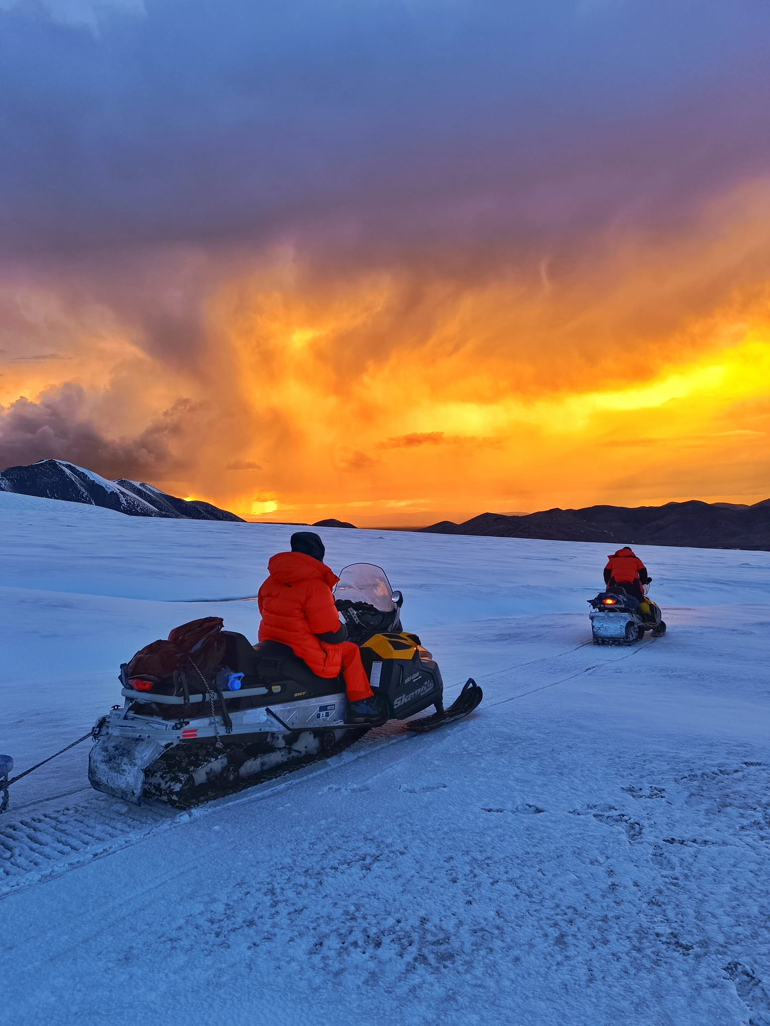 Two people riding two snowmobiles going toward the horizon with orange blue skies and clouds.