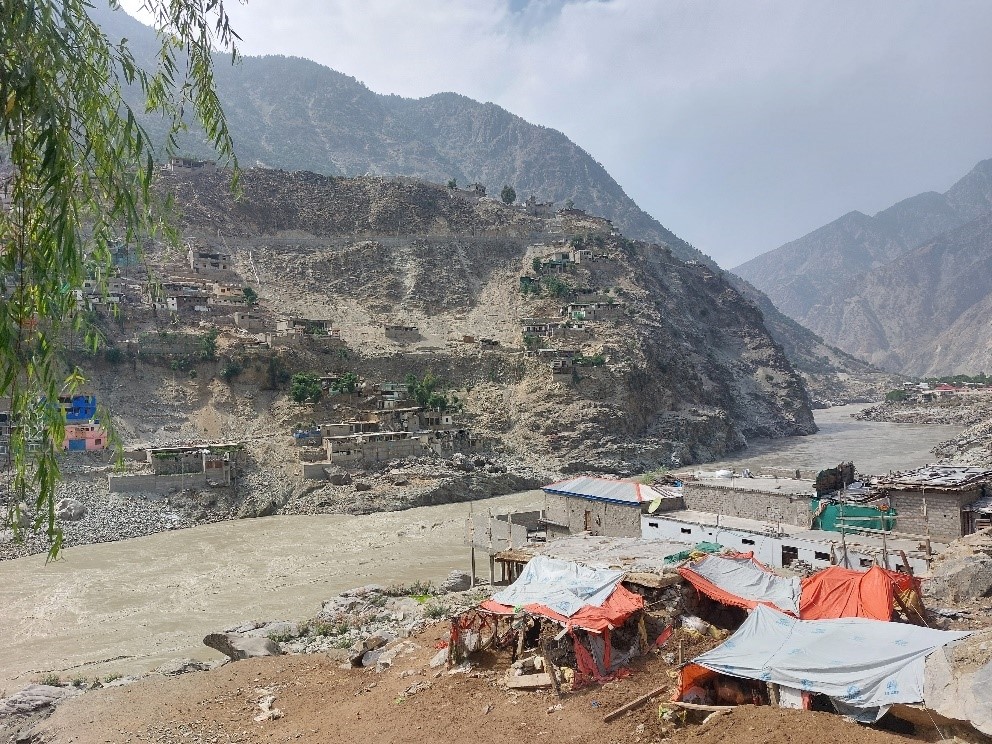 Houses and structures on both sides of the river including tents along the flowing river carved into the hills, with mountains in a distance.