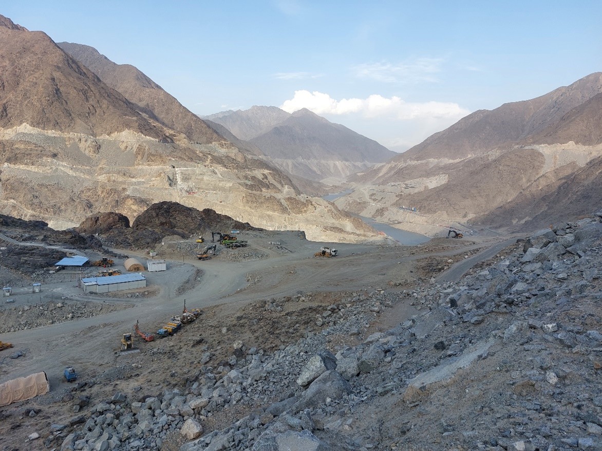 a low lying river in a rocky-dirt terrain in a valley surrounded by brown mountains with some structures, trucks and machinery under blue skies and white clouds.