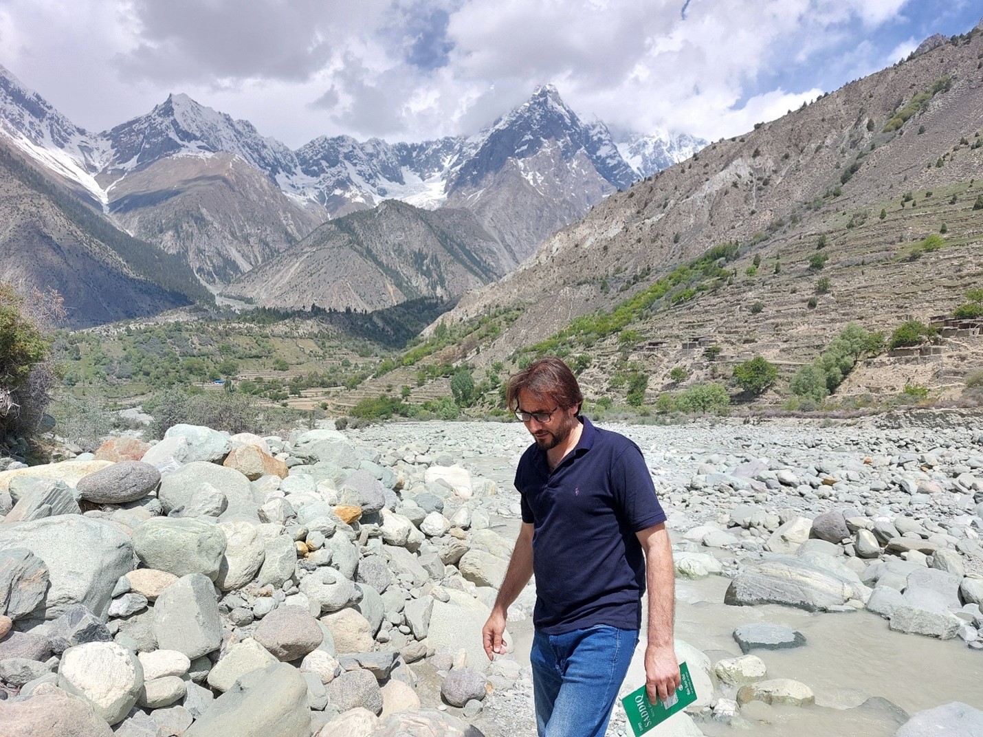 A man holding a green-covered notebook walking in a rocky river bed with a little stream flowing behind him and sparse tree covered hills and mountains in the background. 