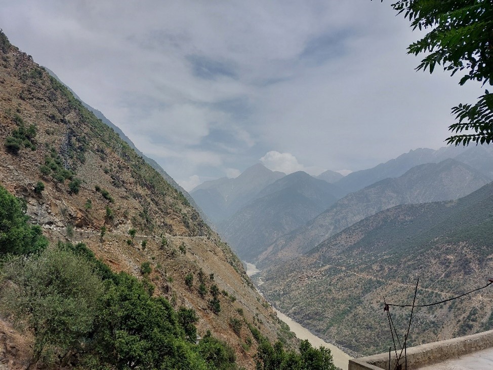 A dirt Mountain range covered sparsely with trees and a river flowing with light brown water below.