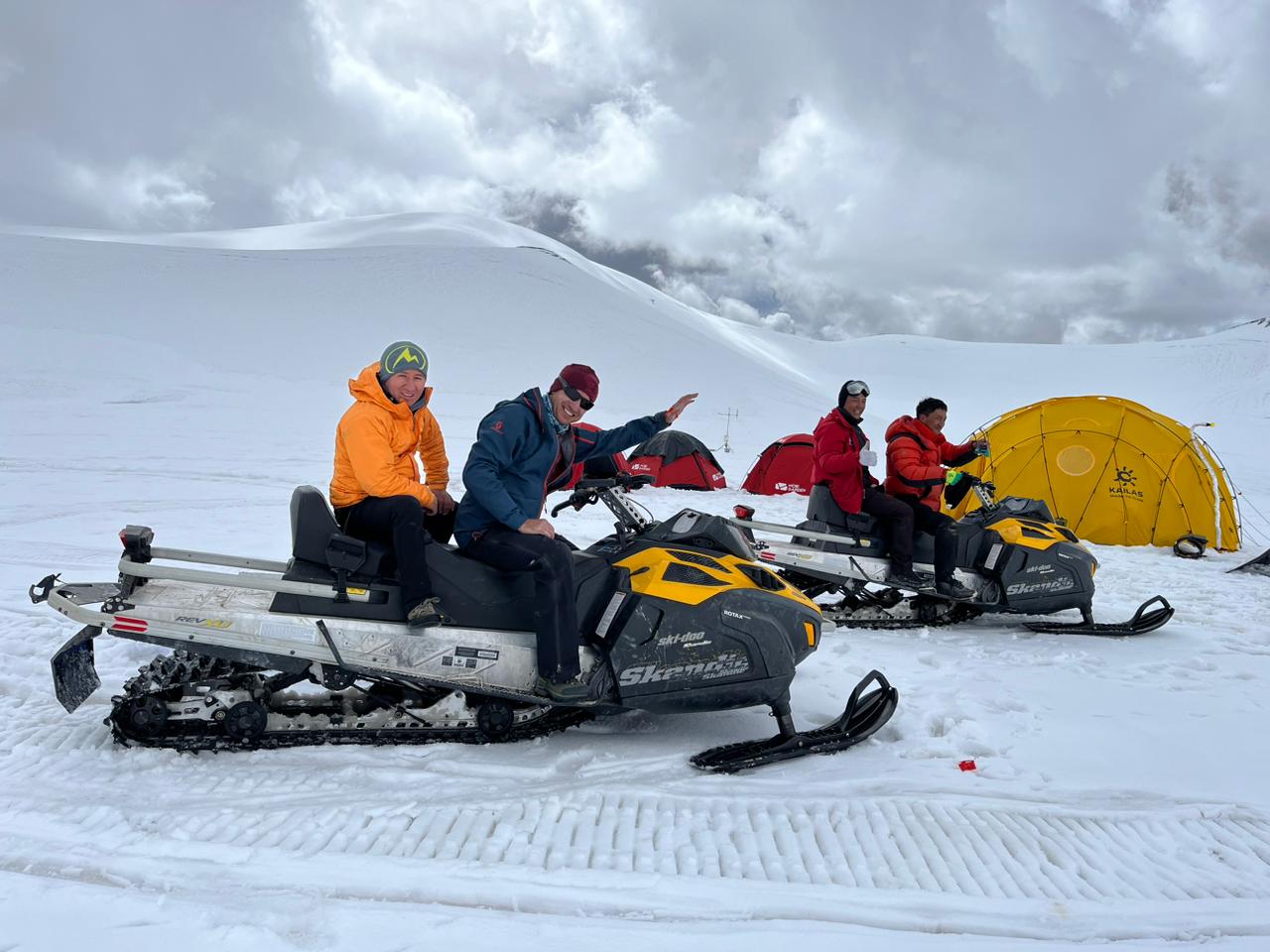 Four people seated on two snowmobiles with red and yellow tents on the snow-covered ground and a snow-covered mountain behind them with white clouds.