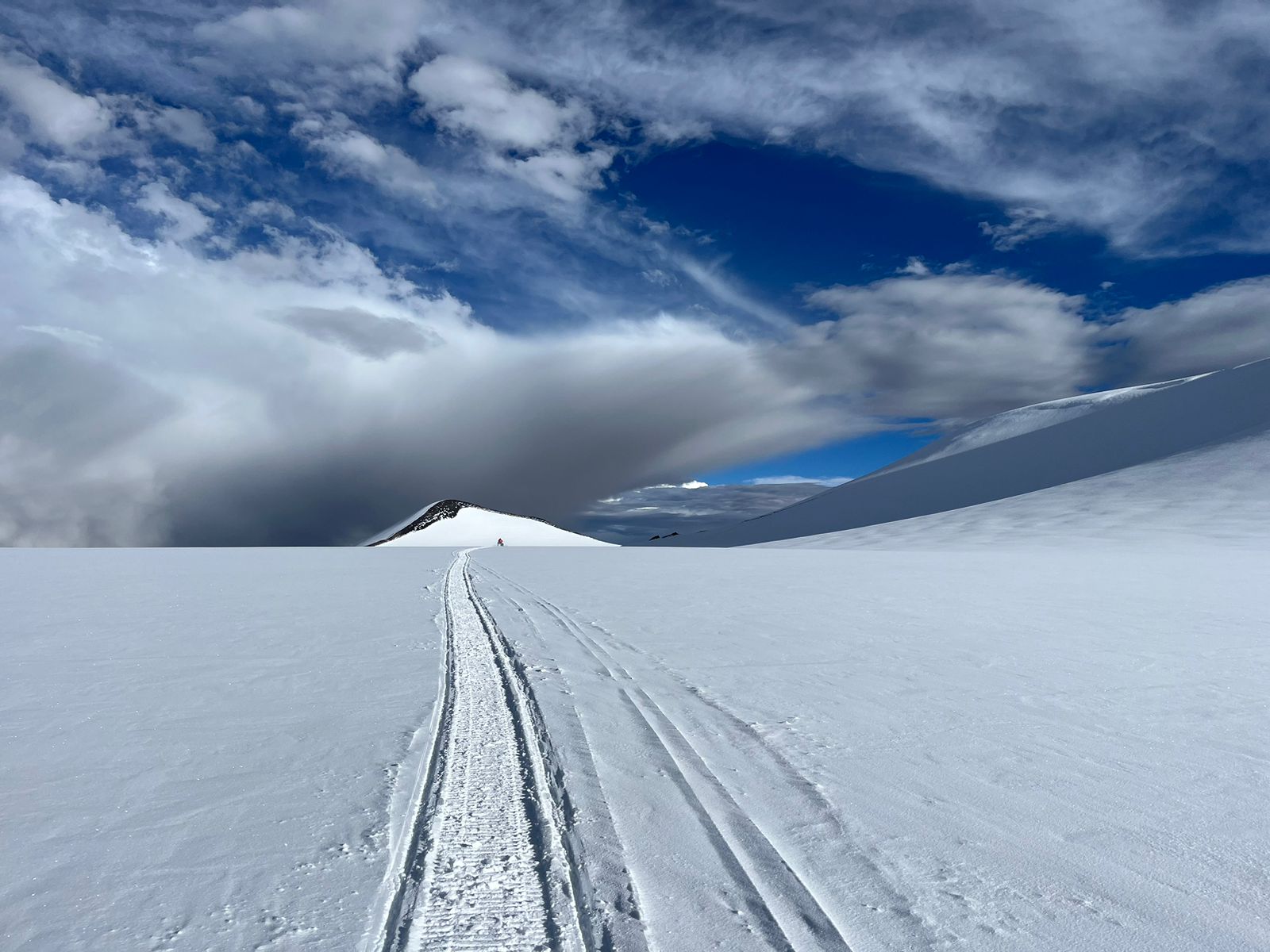 Snow covered ground with snowmobile tracks under blue skies and white clouds.