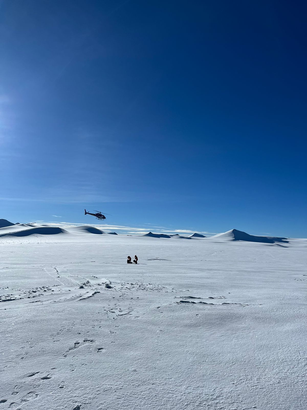 Two People walking in a distance on the snow covered  ground with a vista of mountains and a helicopter flying under blue skies.