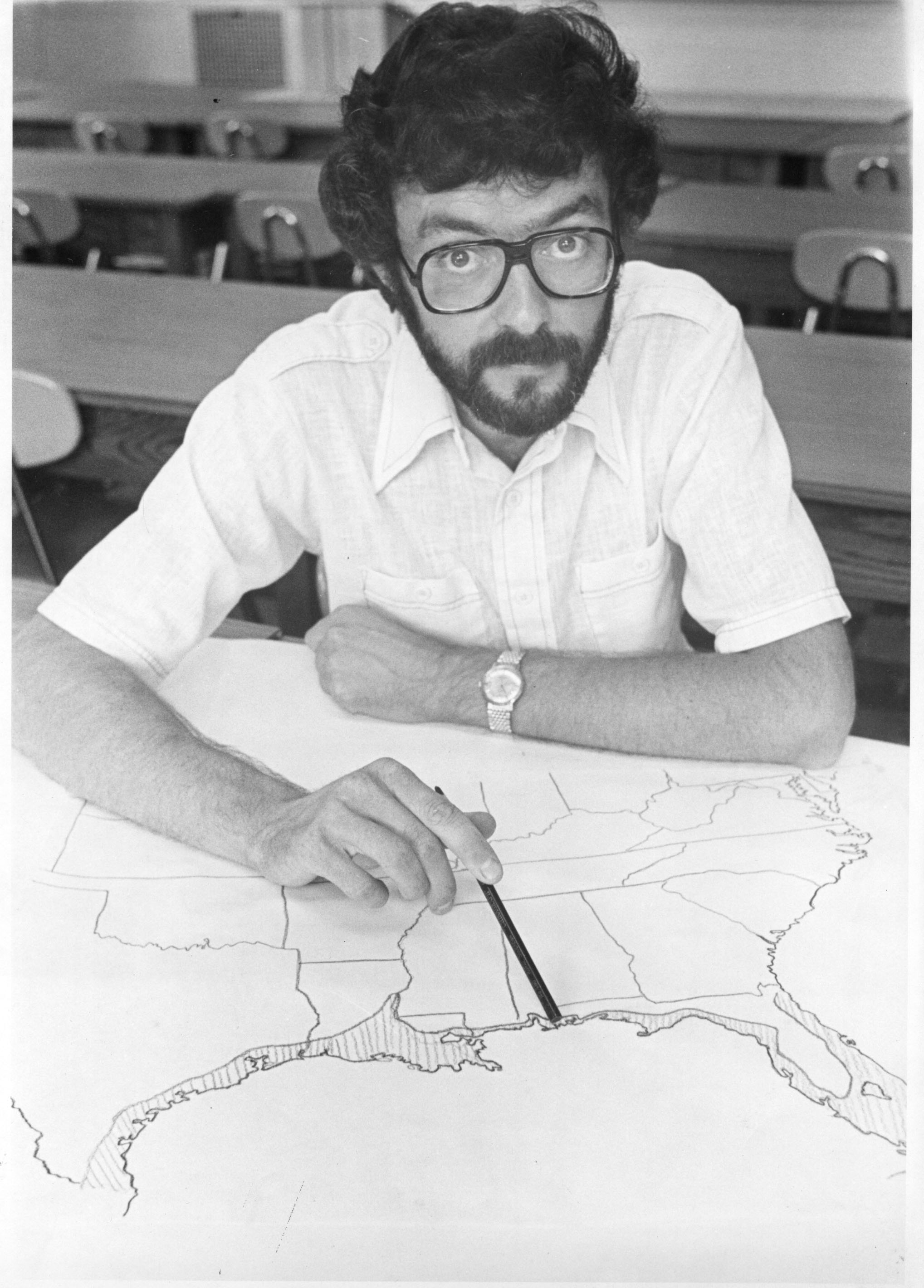 A black and white of a bearded man with glasses sitting in an empty classroom pointing to a map of the US on the table.