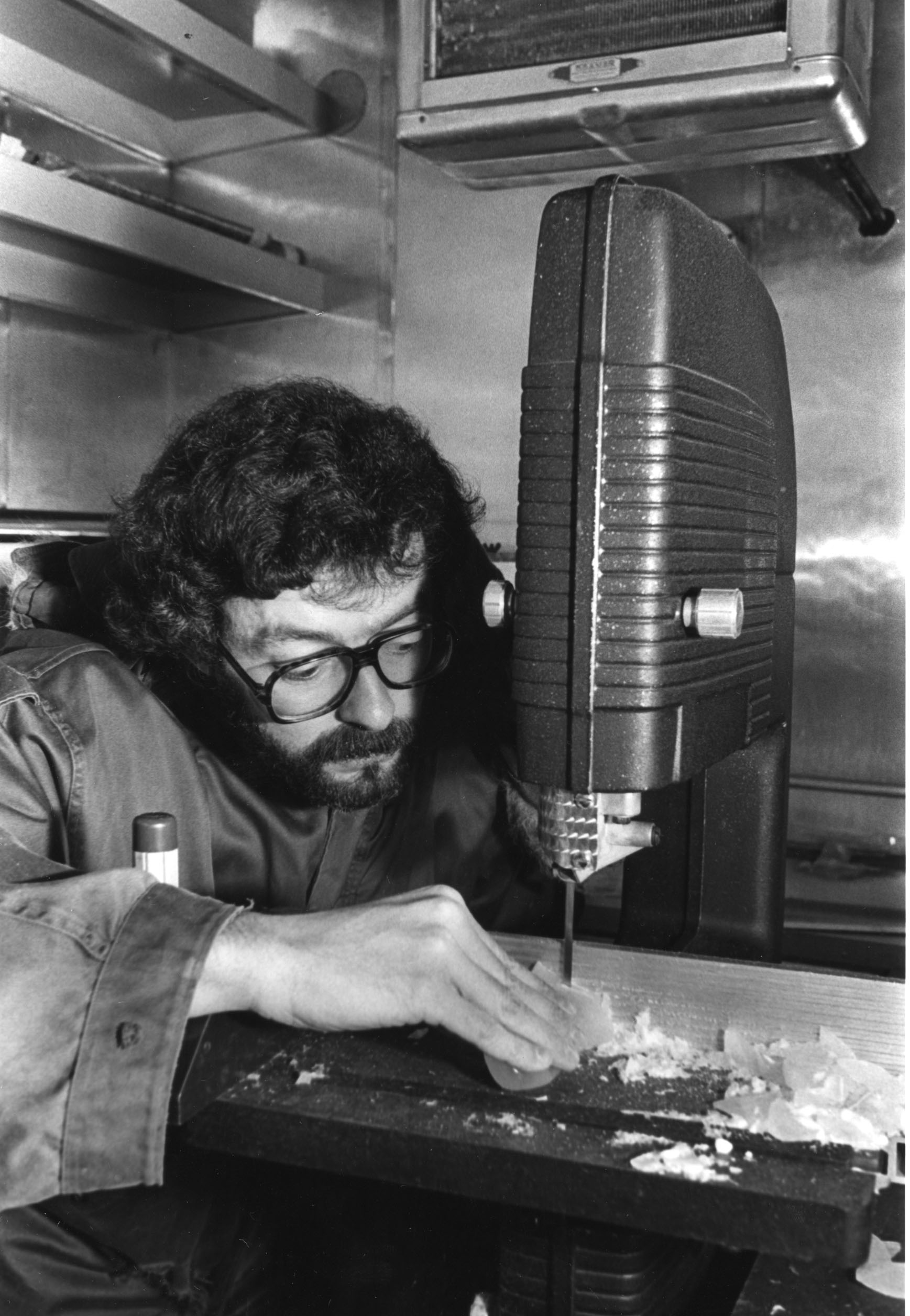 A black and white image of a bearded man with glasses in a winter coat using a table saw to cut ice.