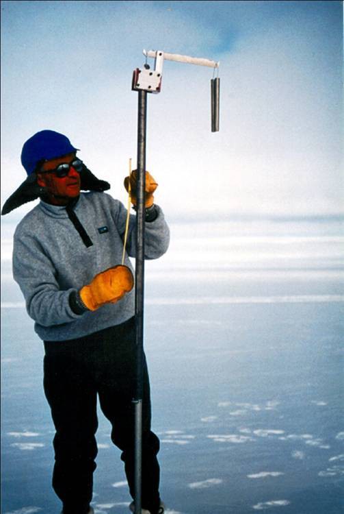 A man standing outdoors, with a snowy background, wearing a winter coat, gloves, sunglasses and hat, holding equipment.