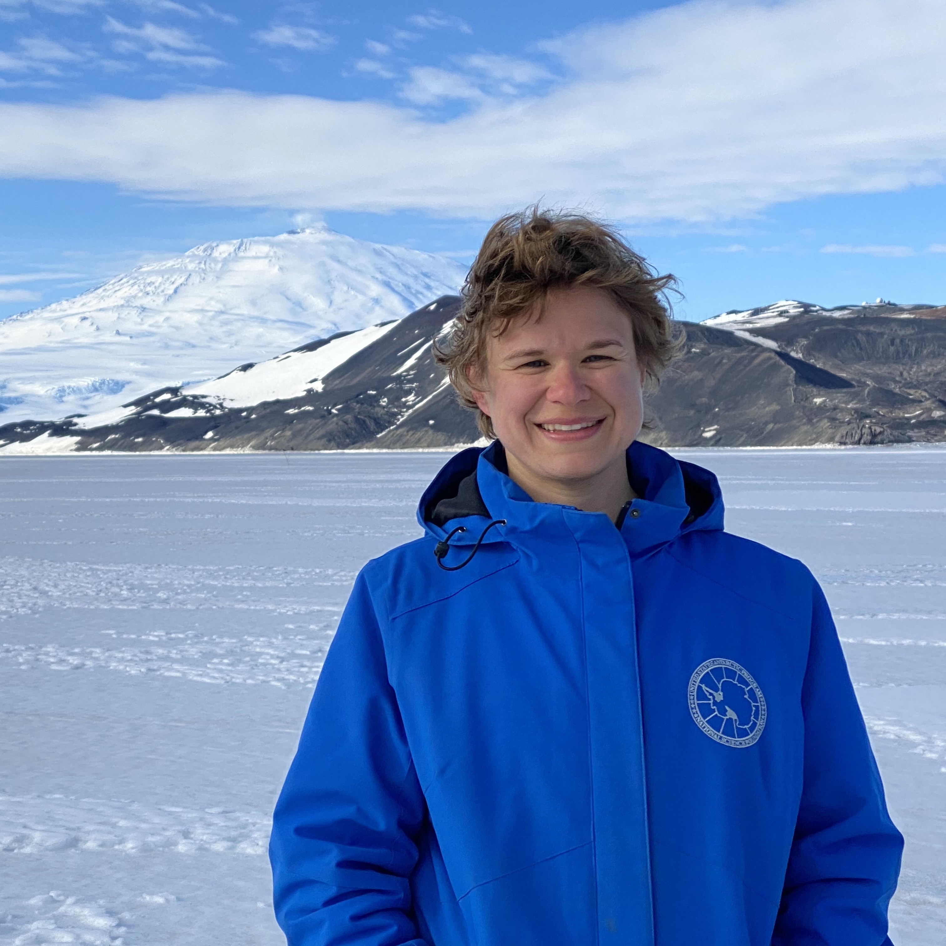 Person in a blue jacket standing in front of a frozen landscape with mountains in the background.