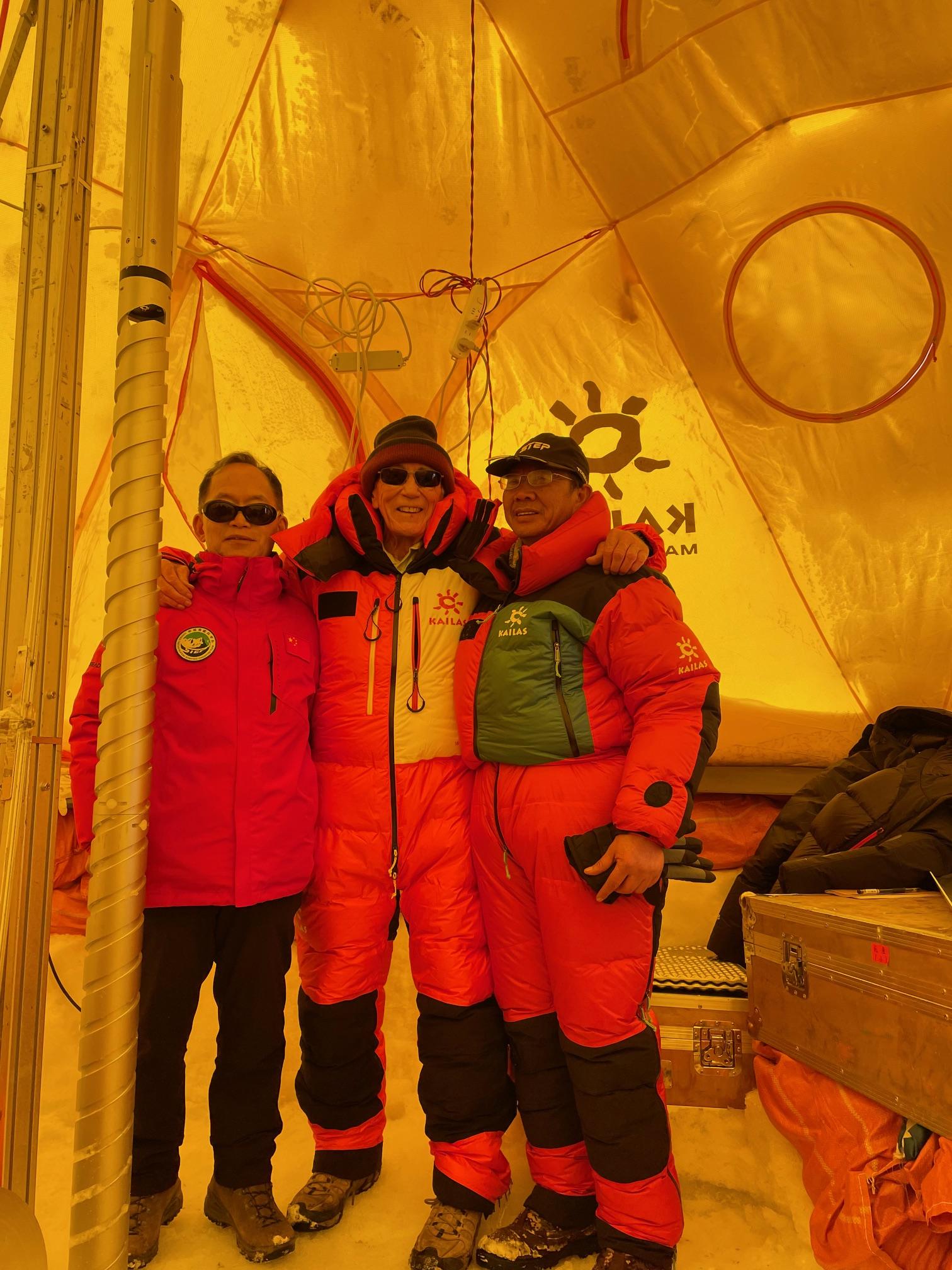 Three climbers in red expedition suits pose inside a tent.