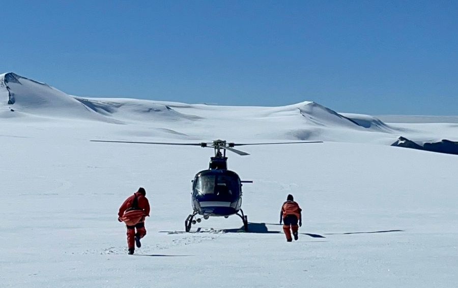 A helicopter and two individuals in red jackets running toward it on a snowy landscape under a clear blue sky.