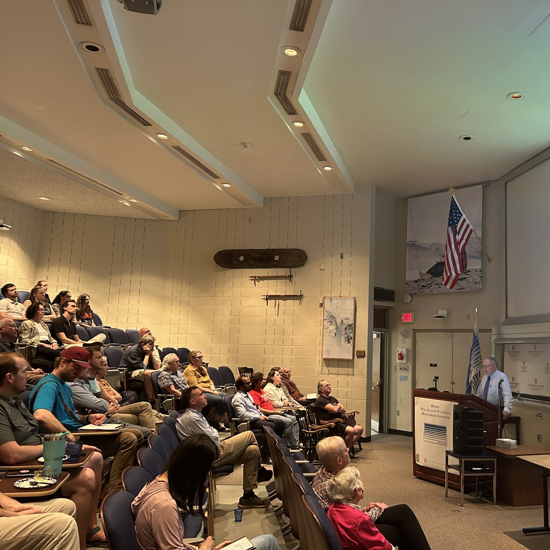 Audience seated in a lecture hall listening to a speaker at a podium, with a canoe mounted on the side wall.