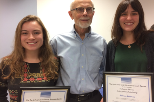 Three individuals pose indoors against a plain background. The two individuals on either side are holding certificates.