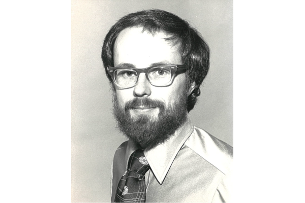Black and white portrait of a man with glasses, a full beard, and short hair, wearing a collared shirt and a tie.