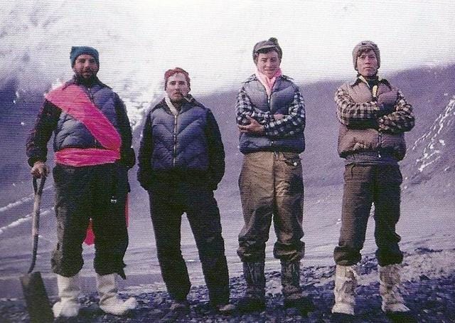 Four men standing outdoors, the one on the left holding a shovel, in winter clothes with a backdrop of snow covered mountains.  
