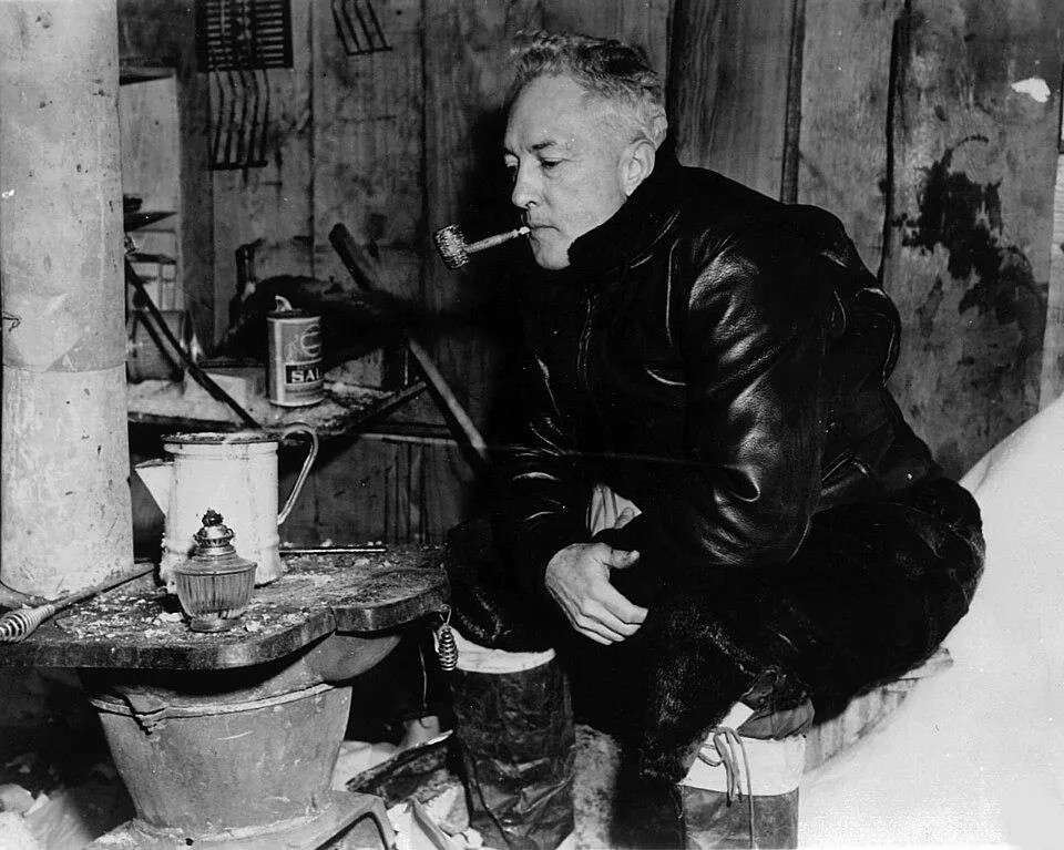 A black and white of a man in winter gear smoking a pipe indoors with supplies around hom.