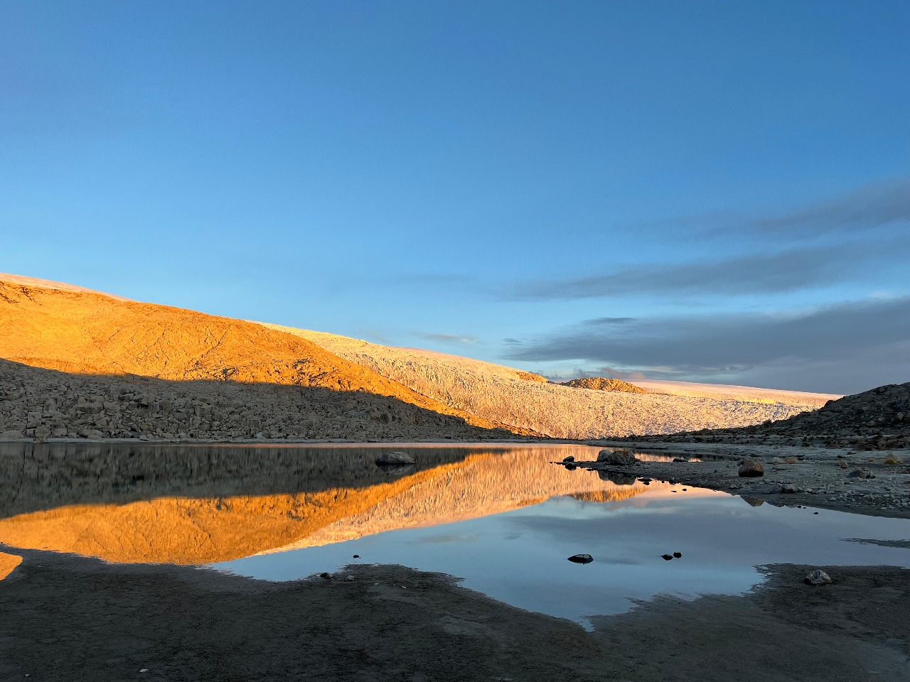 Sunrise reflects golden light on a rocky mountain slope across a still lake.