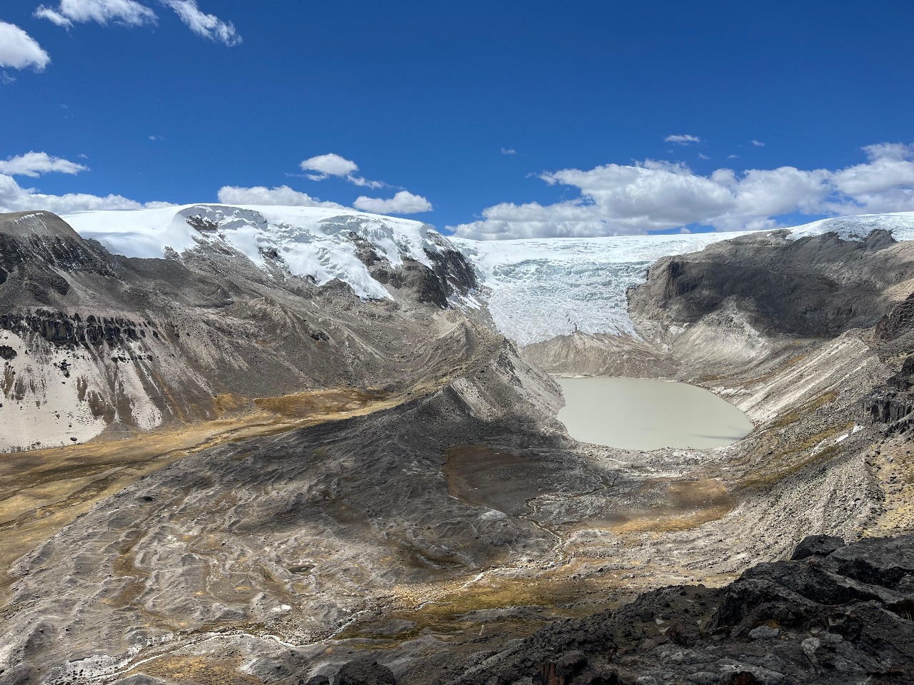 Snow-capped glacier descends into a gray lake surrounded by rocky mountains under a bright blue sky.