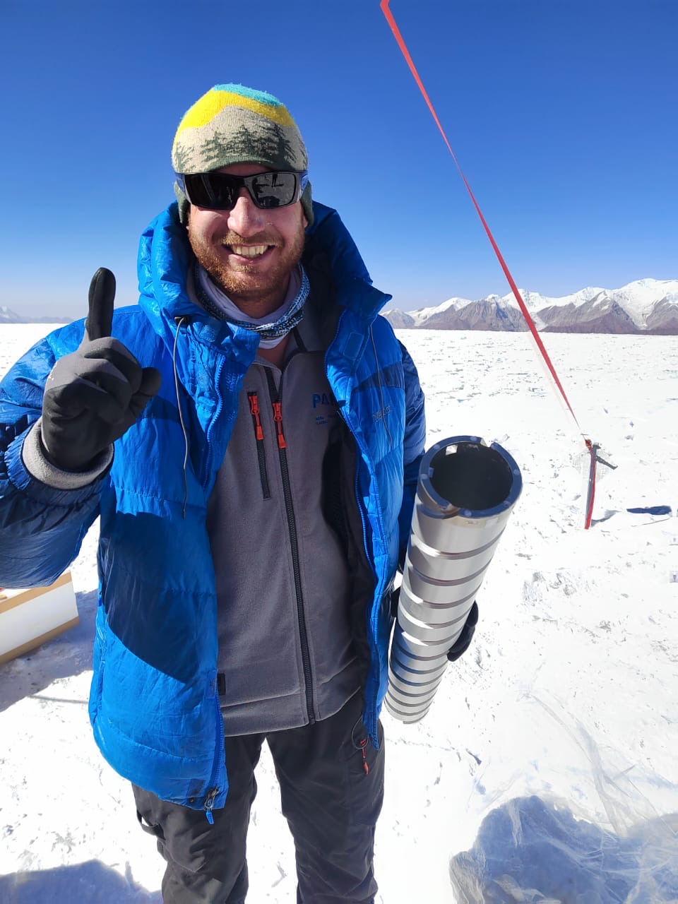 Smiling researcher on snowy glacier holding an empty ice-core barrel.