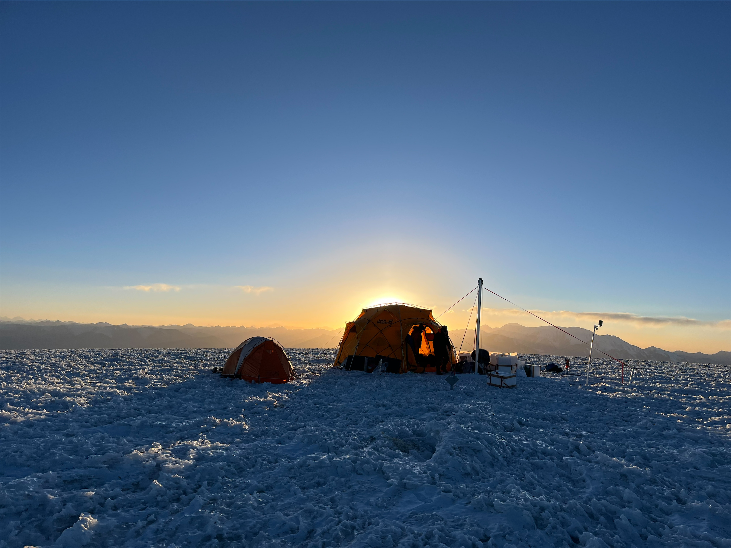 Glacier-top camp with lit tents under low-angle light near the horizon.