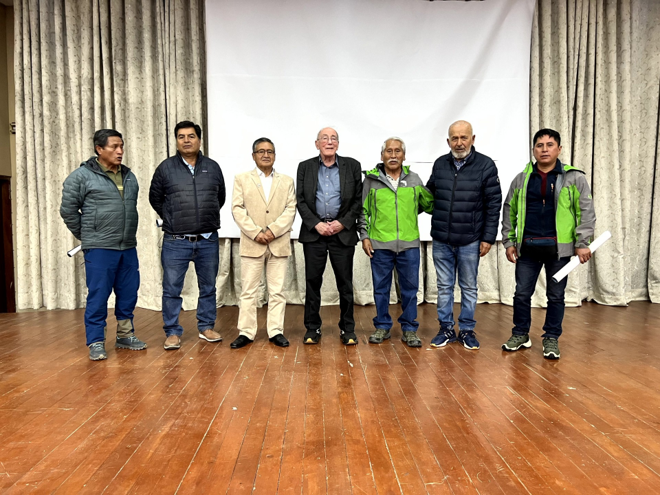 Seven men stand side by side on a stage in front of a curtain and projection screen, posing for a group photo.
