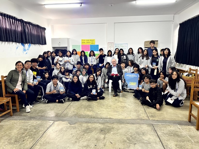 Group photo of high school students and teachers gathered around a seated guest speaker holding a CANARY movie poster.