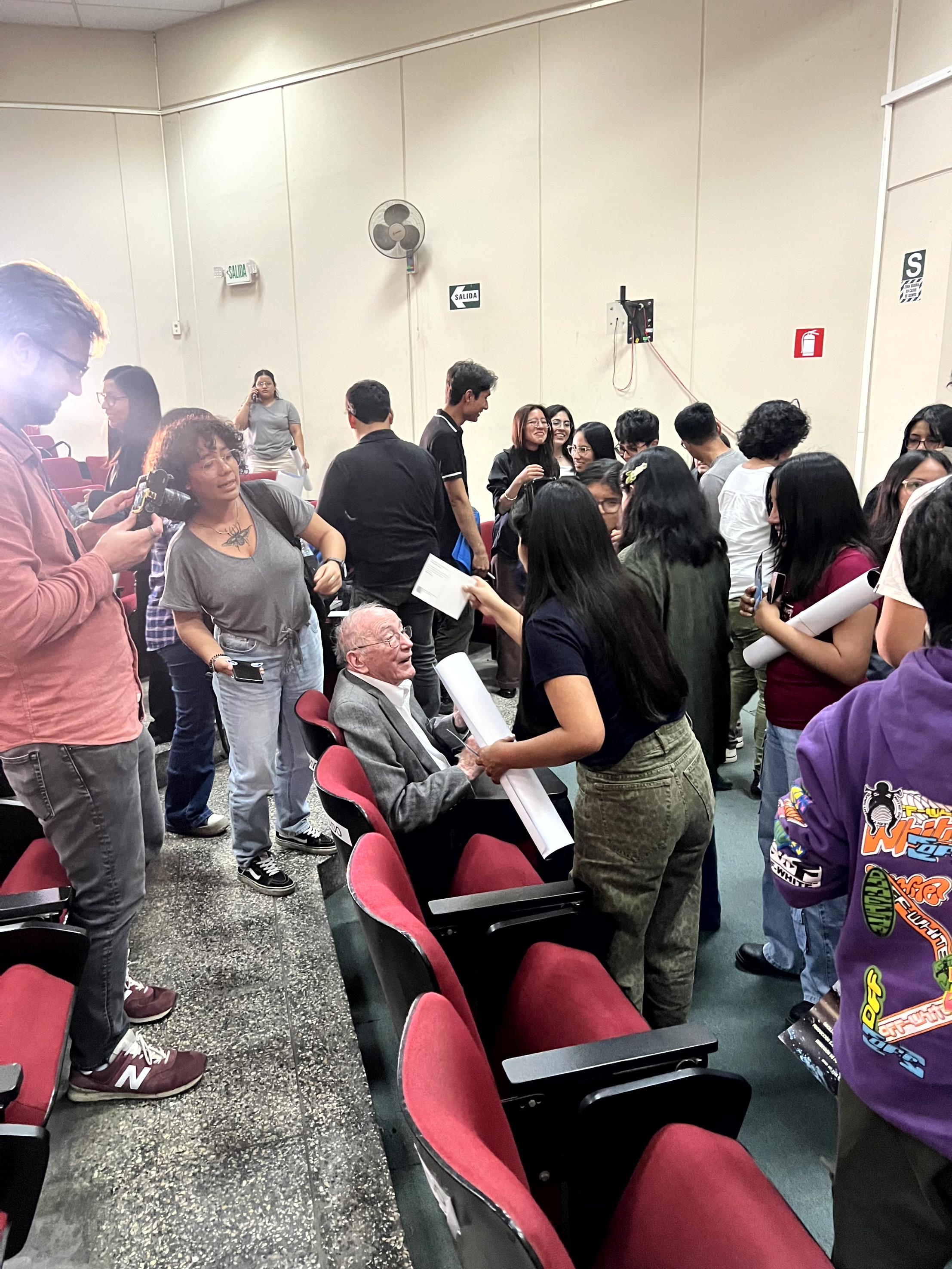 people gather around an older man seated in an auditorium, chatting and holding papers after an event.