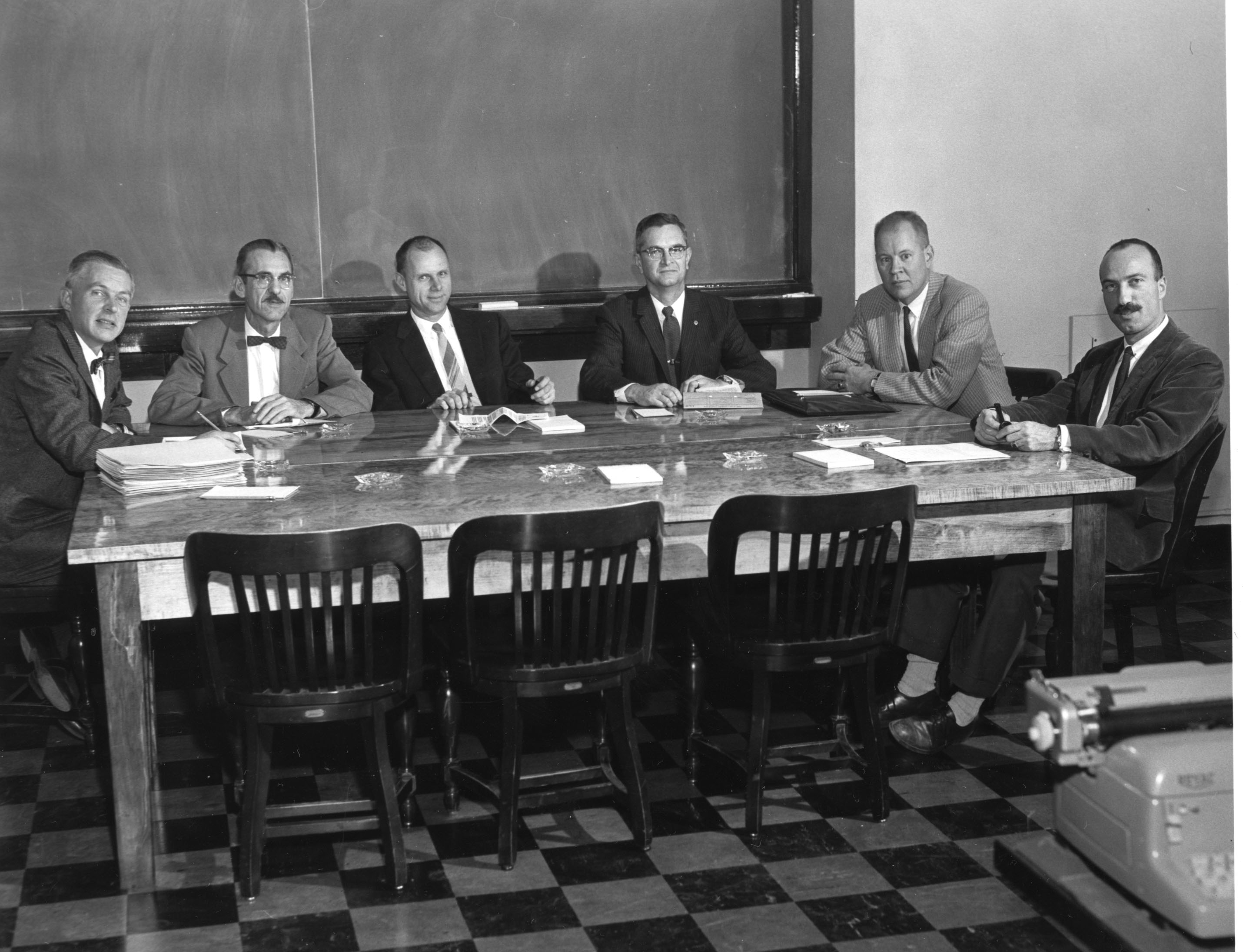 A black and white photo of a group of men in suits sitting around a table with many documents strewn about.