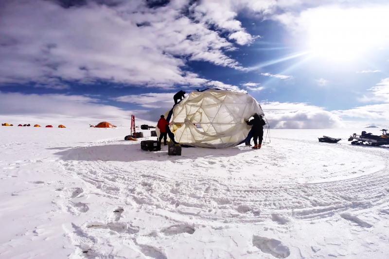 Two people setting up a dome tent under blue, sunny skies with white clouds.