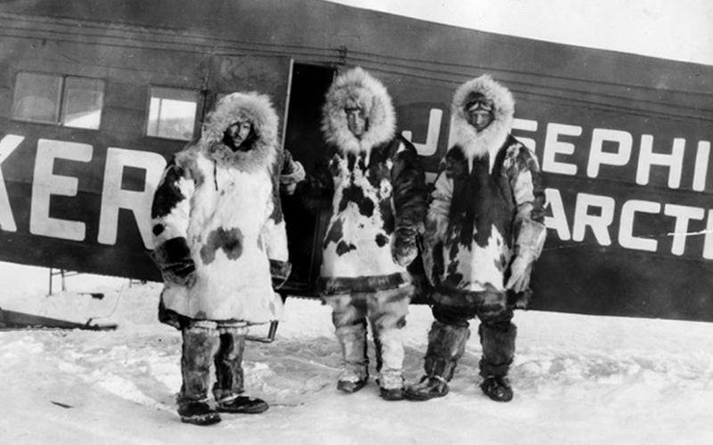 a black and white photo of three people dressed in thick coats with furlined hoods. They are standing in the snow. Behind them is a plane.