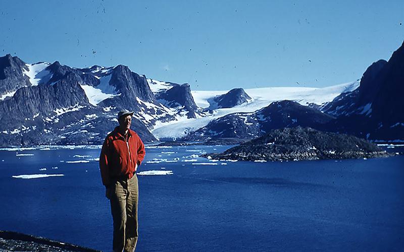 A man poses in a red jacket in front of a snowy, rocky mountain range behind a body of water 