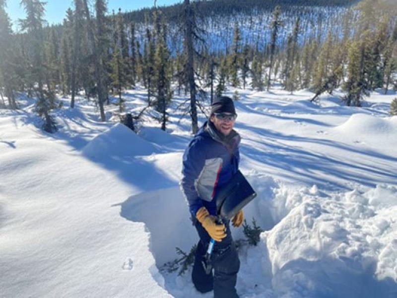 Michael Durand digging a snowpit in Alaska in field of snow and some pine trees in background