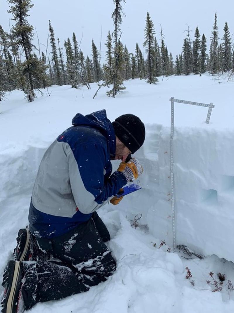Dr. Durand on the ground looking through equipment at snow in Alaska in a landscape of snow and trees