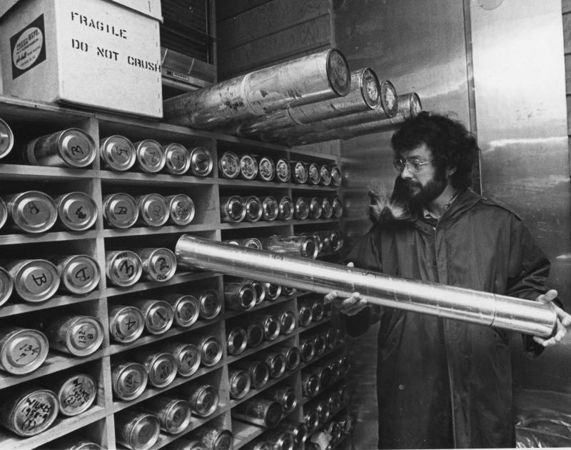 A black and white image of a bearded man with glasses in a winter coat holding a silver cylinder, against a shelf filled with cylinders along the wall.