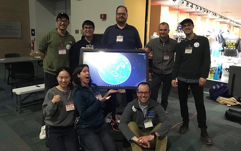 A group of people pose for a photo on front of a screen displaying their work