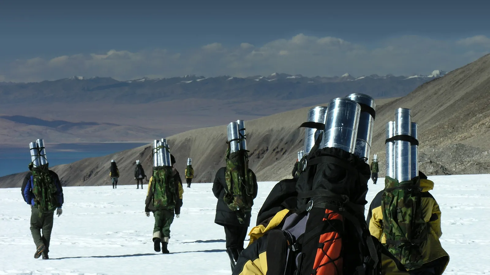 A group of people walking on a snowy ground and brown mountains in a distance, with silver cylinders on their backs.
