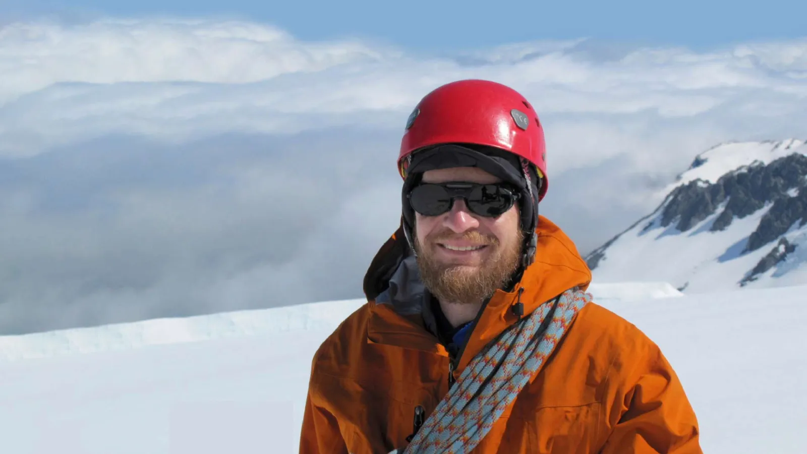 Richard Alley standing on a snow covered mountain. Richard is wearing a helmet and an orange coat with a length of rope hung across his chest